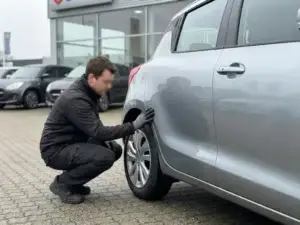 Persoon die zorgvuldig de carrosserie en deurpanelen van een zilvergrijze Suzuki Swift inspecteert bij een autodealer.