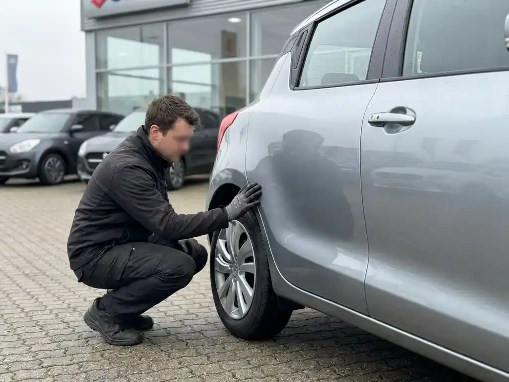 Persoon die zorgvuldig de carrosserie en deurpanelen van een zilvergrijze Suzuki Swift inspecteert bij een autodealer.