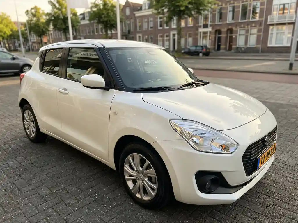 Nauwelijks gebruikte Suzuki Swift op een autodealer in Den Haag, glanzende witte lak en zichtbare kilometerstand door de voorruit.
